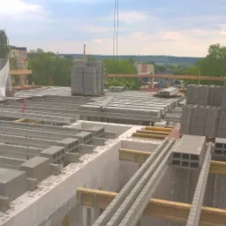 Wznoszenie stropu Teriva na budowie domu jednorodzinnego, widoczne belki stropowe, pustaki, podesty robocze i fragment panoramy miasta w tle.