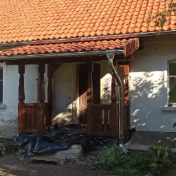 Dom z pomarańczową dachówką, widoczne nowe rynny i odnowione drewniane elementy werandy, teren przygotowany do dalszych prac remontowych.
