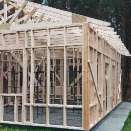 Konstrukcja drewniana szkieletowa domu jednorodzinnego w trakcie budowy, widoczne elementy dachu i ścian, otoczenie leśne.