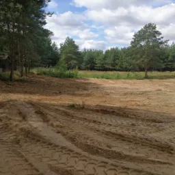 Wyrównany teren w Ostrowite, przygotowany pod inwestycję, widoczne ślady opon ciężkiego sprzętu, w tle las iglasty i błękitne niebo z chmurami.