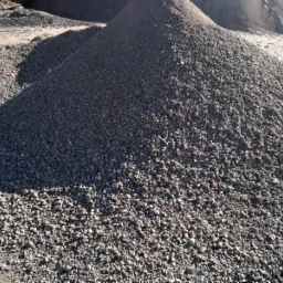 Góra drobnego, szarego kruszywa na placu budowy, częściowo zacieniona, z fragmentem opony pojazdu w tle.