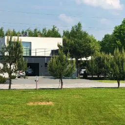 Nowoczesny dom jednorodzinny z płaskim dachem, szarą elewacją i balkonem, otoczony zielenią, z dwoma samochodami zaparkowanymi w garażu.