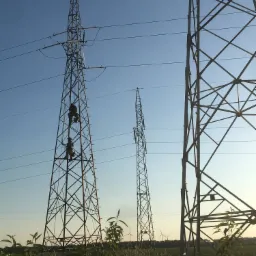 Dwóch pracowników na stalowej wieży wysokiego napięcia w okolicach Poznania, wykonujących prace konserwacyjne na wysokości, widok z dołu na tle błękitnego nieba i polnej roślinności.