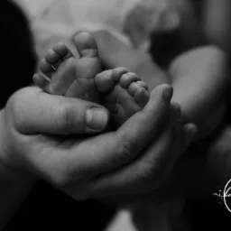Drobne stopy noworodka delikatnie trzymane w dłoni rodzica, czarno-biała fotografia z logo fotografa w prawym dolnym rogu.