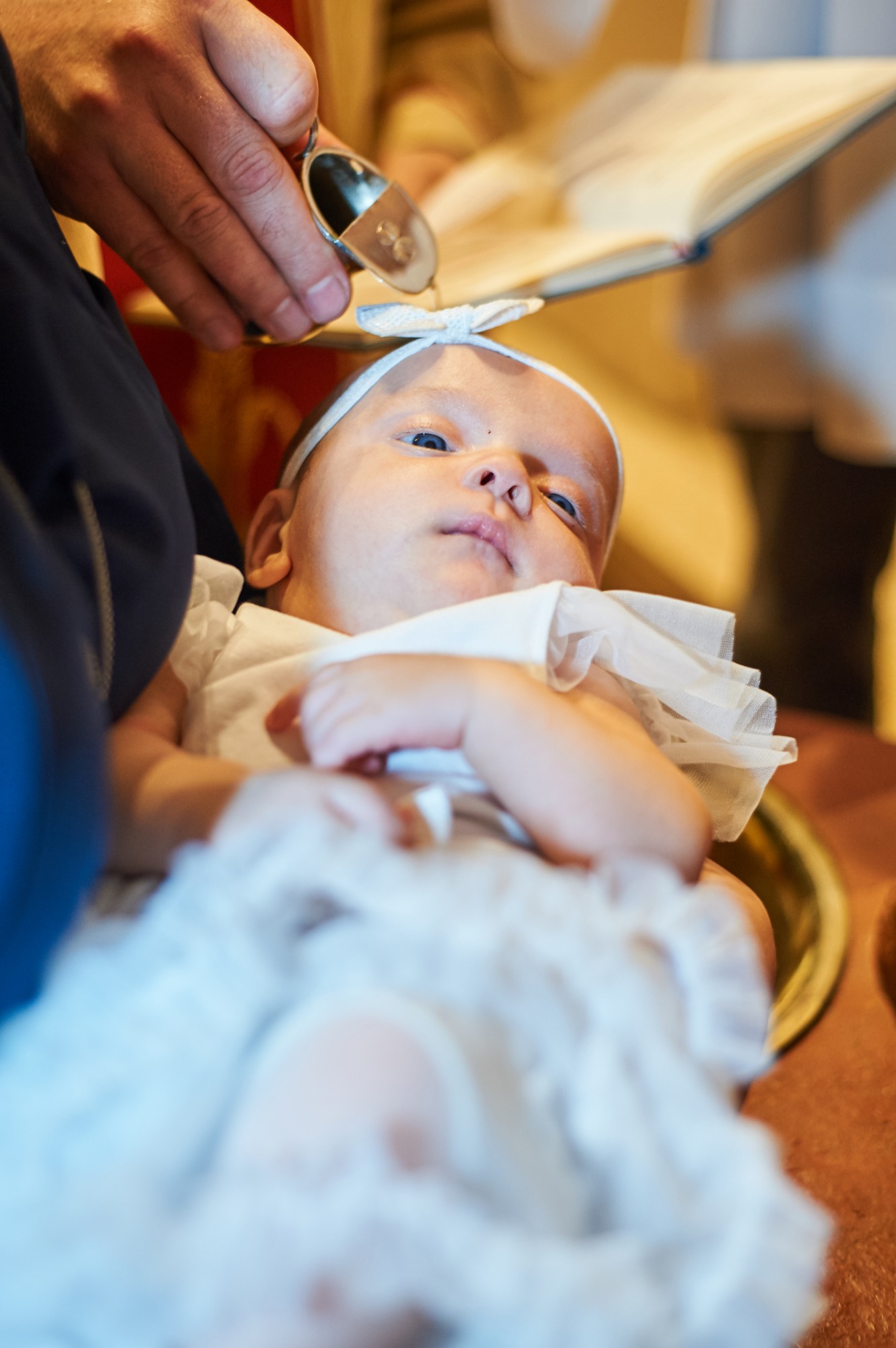 Niemowlę z niebieskimi oczami podczas chrztu, w tle księga i dłoń kapłana z naczyniem na wodę.