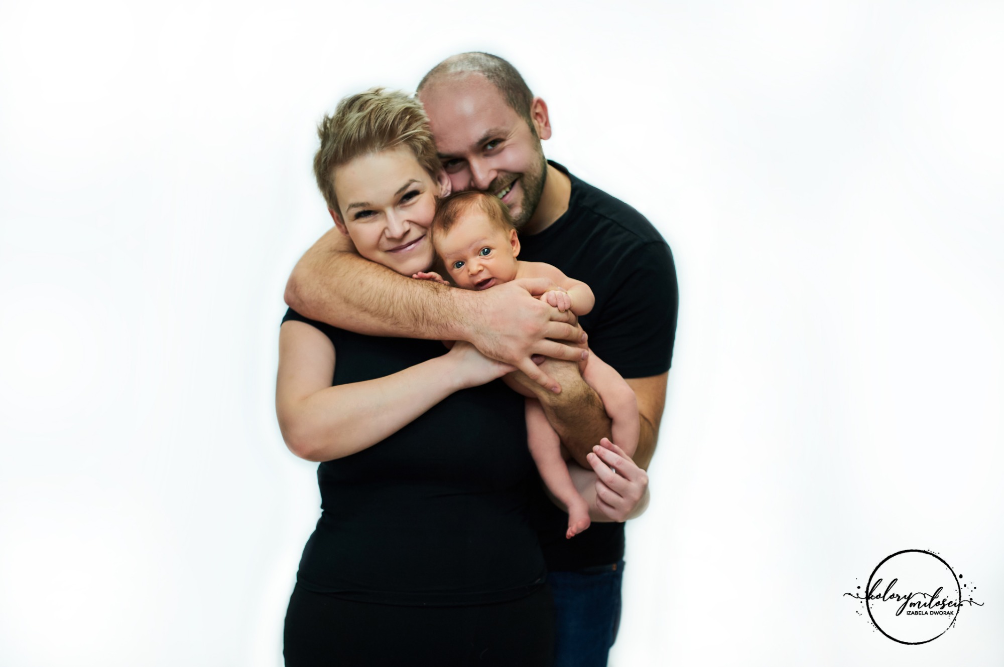 Rodzinne zdjęcie studyjne: uśmiechnięci rodzice w czarnych ubraniach trzymają nagiego noworodka na białym tle, widoczne logo fotografa w prawym dolnym rogu.