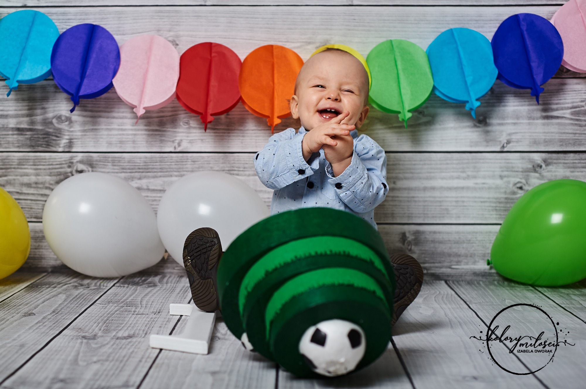 Uśmiechnięty roczny chłopiec w błękitnej koszuli siedzi na drewnianej podłodze z kolorowymi papierowymi balonami w tle, białe i zielone balony po bokach, a przed nim duża, zielona zabawka...