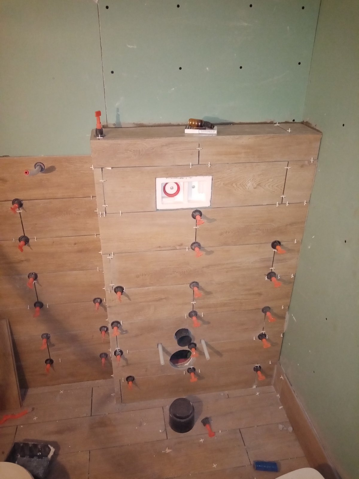 Układanie drewnopodobnych płytek na ścianie z widocznymi otworami na rury i stelaż podtynkowy, używając poziomujących klipsów i krzyżyków dystansowych.