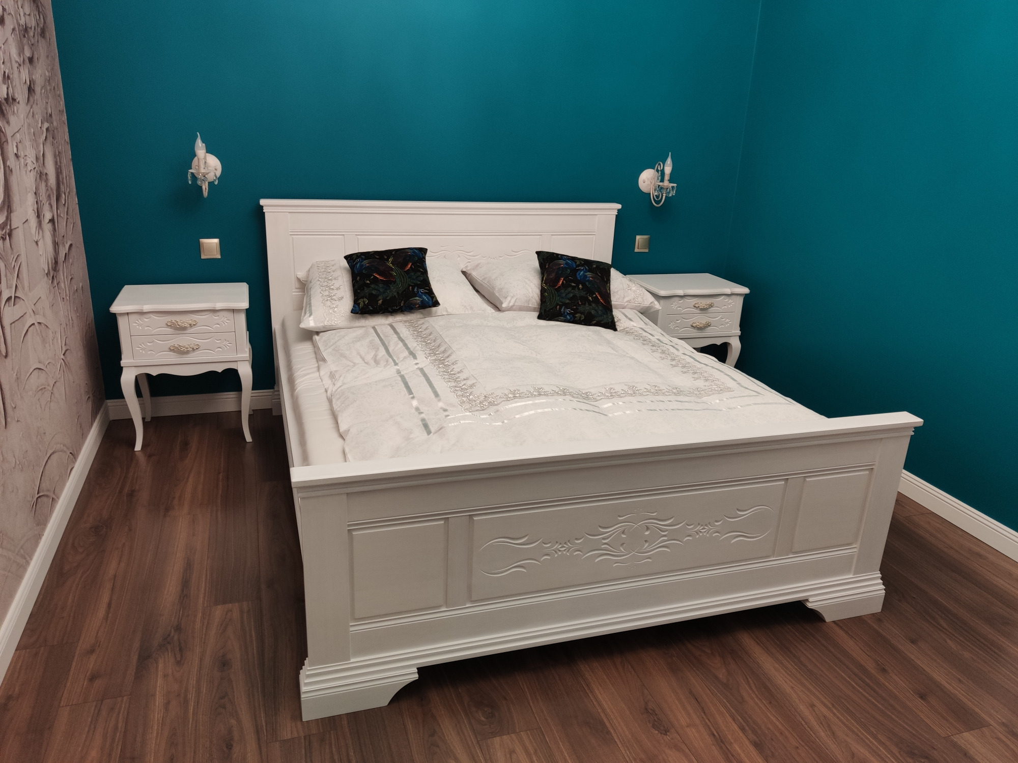 Białe łóżko z dekoracyjnymi rzeźbieniami i szafki nocne w pokoju z turkusowymi ścianami i drewnianą podłogą.