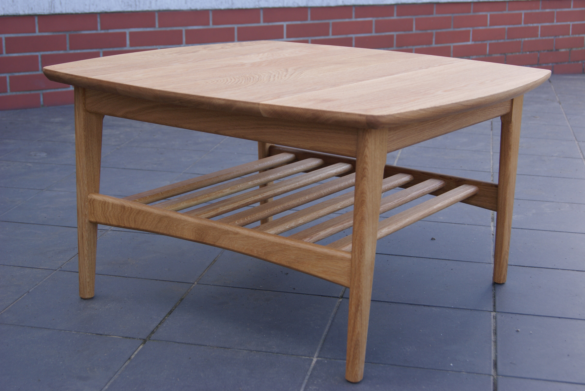 Stolik kawowy z jasnego drewna dębowego, z półką z listewek, na tle ściany z czerwonej cegły i szarej terakoty.