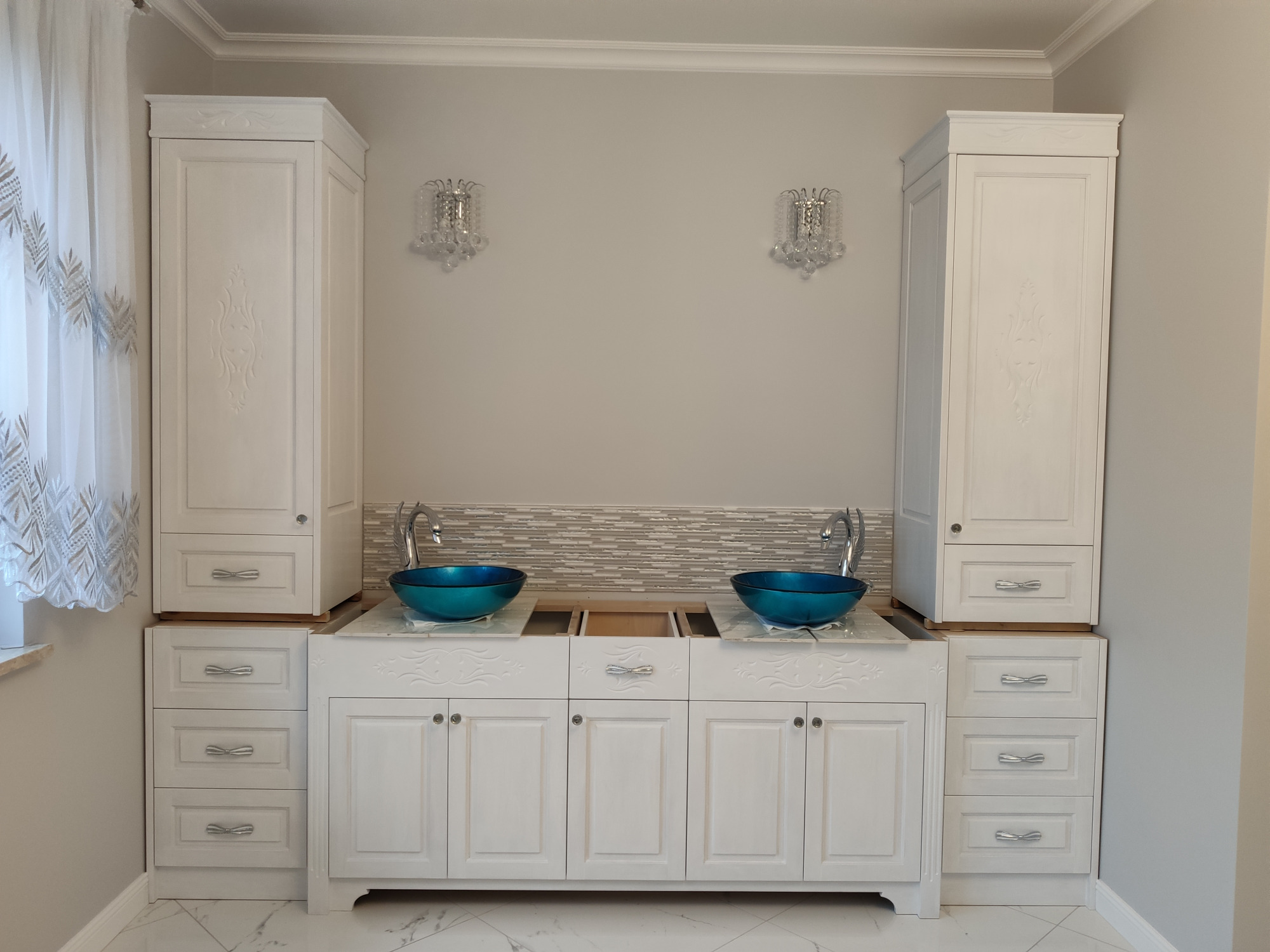Biała, symetryczna zabudowa łazienkowa z dwiema szafkami wiszącymi, szafkami dolnymi z szufladami i dwoma turkusowymi umywalkami na blacie z imitacją marmuru, widoczne stylowe kryształowe kinkiety.