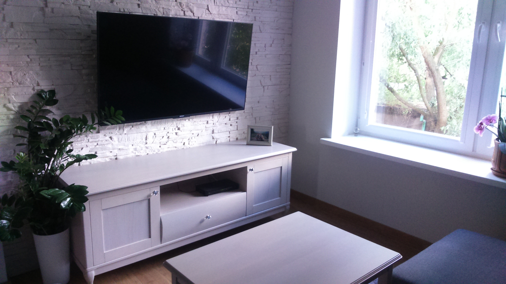 Jasna szafka RTV z szufladą i półką pod telewizorem na ścianie z imitacji kamienia, obok okno z widokiem na drzewo.