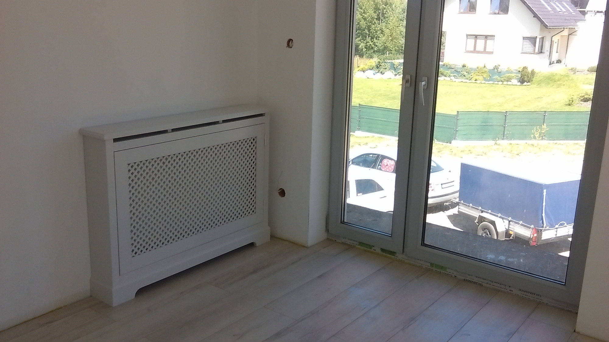 Biała, ażurowa obudowa grzejnika z płyty MDF, stojąca w rogu pokoju z jasnymi panelami podłogowymi, na tle okna z widokiem na dom, trawnik i przyczepkę samochodową.