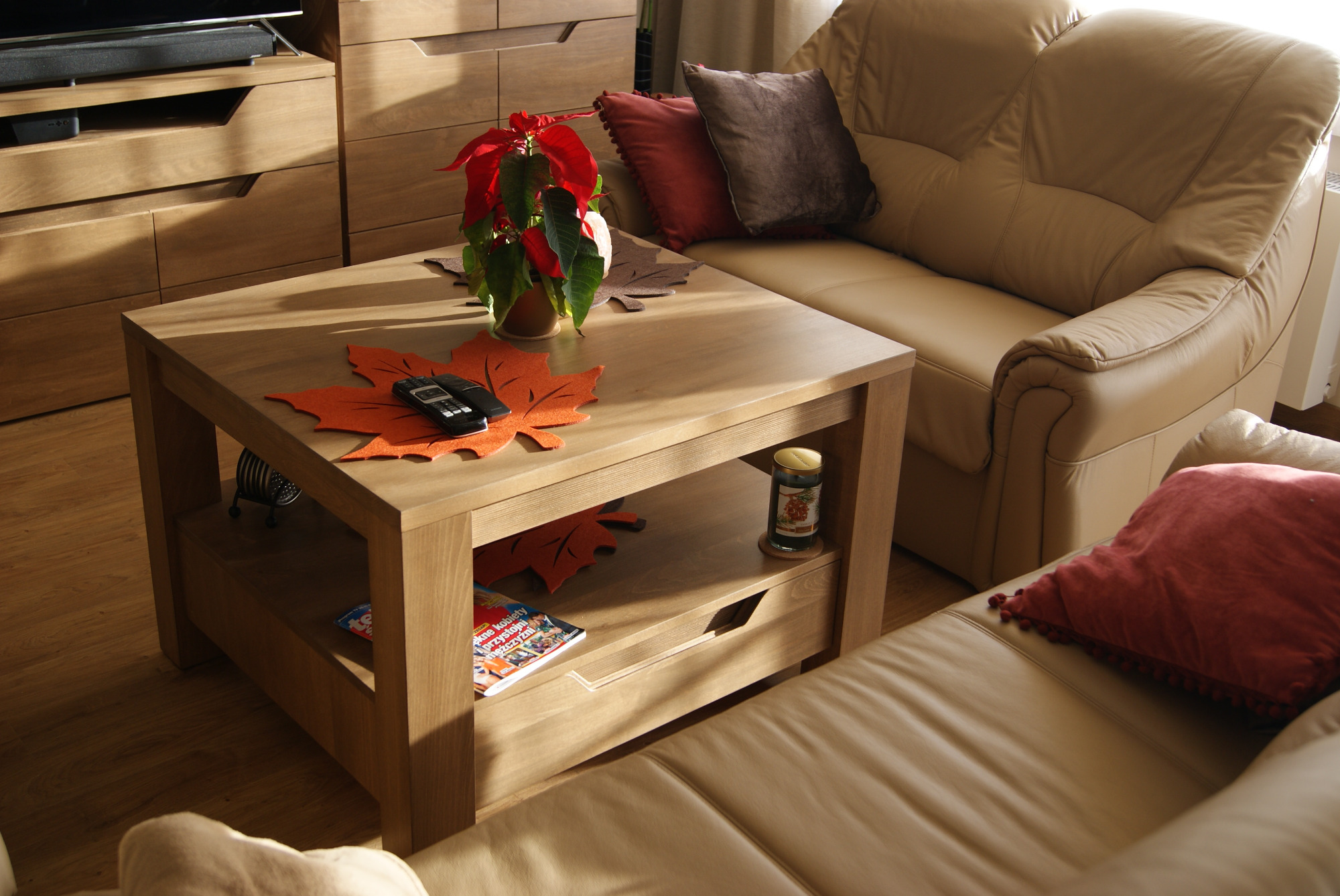 Jasny salon z drewnianymi meblami: stolik kawowy z półką, szafka RTV i tapicerowany narożnik w beżowej skórze, dekoracje w postaci czerwonej gwiazdy betlejemskiej i poduszek.