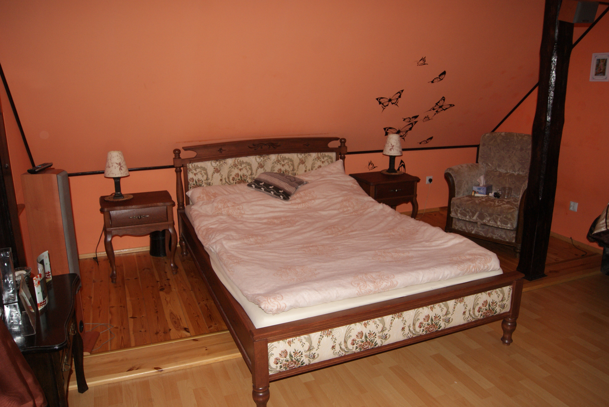 Pokój na poddaszu z łóżkiem z tapicerowanym dołem w kwiatowy wzór, drewnianymi szafkami nocnymi i fotel z poduszkami, ściany w kolorze pomarańczowym zdobione motylami.