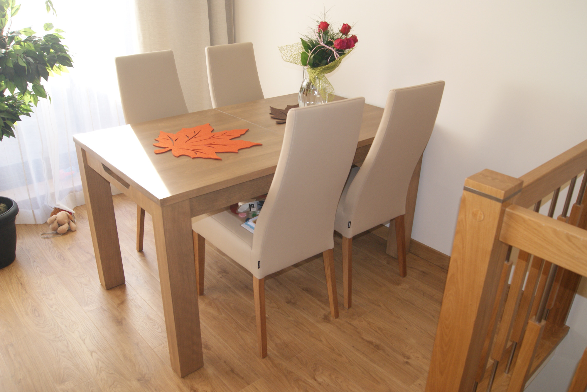 Drewniany stół z czterema krzesłami o jasnej tapicerce, na blacie dekoracja w kształcie liścia klonu i wazon z różami, w tle widoczny fragment schodów.