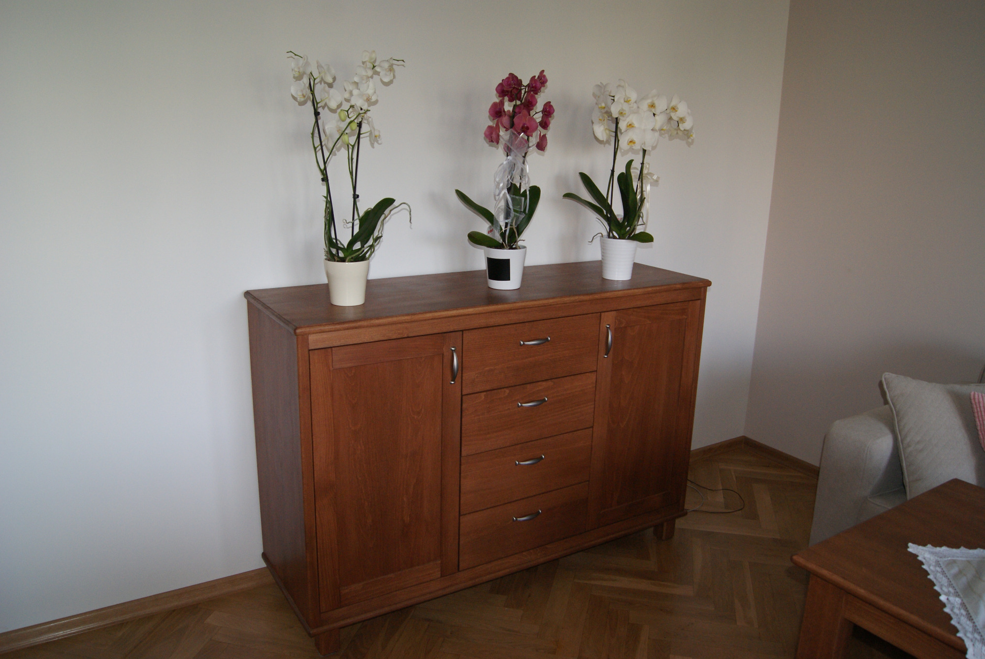 Drewniana komoda z trzema szufladami i dwojgiem drzwiczek, na blacie trzy doniczki z kwitnącymi storczykami, ustawiona na parkiecie w jasnym wnętrzu.
