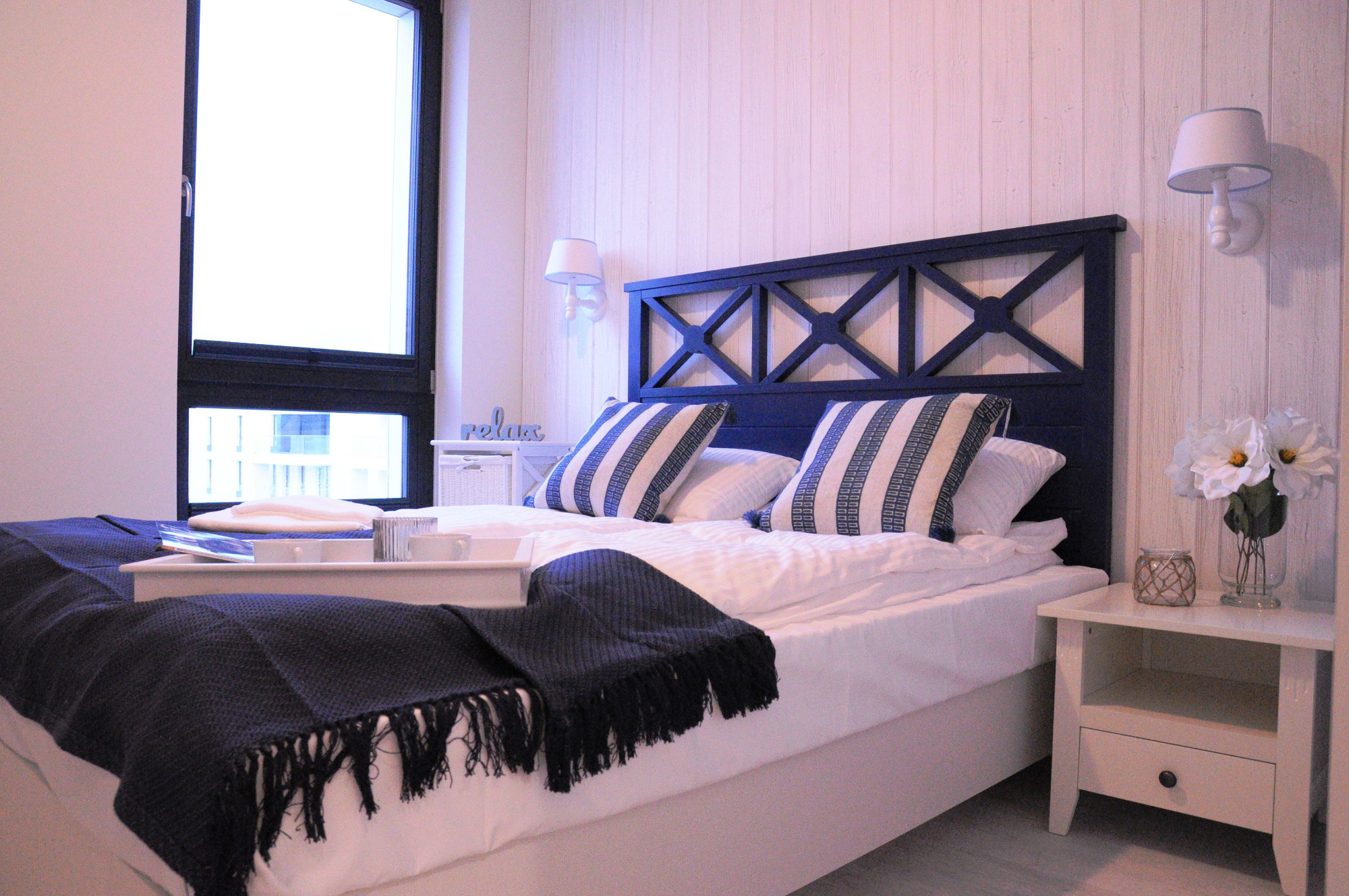 Sypialnia w stylu marynistycznym z ciemnoniebieskim zagłówkiem łóżka z motywem krzyży, białą pościelą w paski, stolikami nocnymi i dekoracjami, oświetlona światłem dziennym wpadającym przez okno.