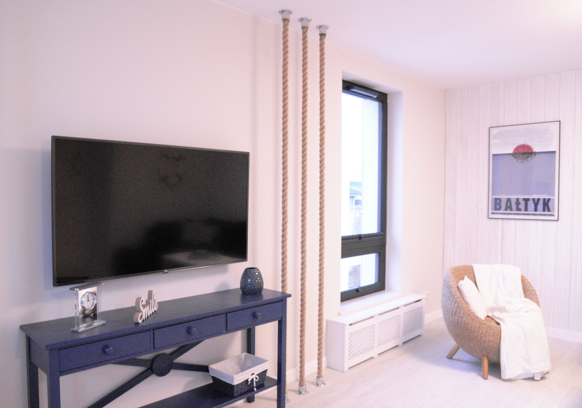 Jasne wnętrze pokoju w Wierzchowie z ciemnoniebieską konsolą pod telewizorem, wiklinowym fotelem z kocem i dekoracyjnymi linami od podłogi do sufitu.