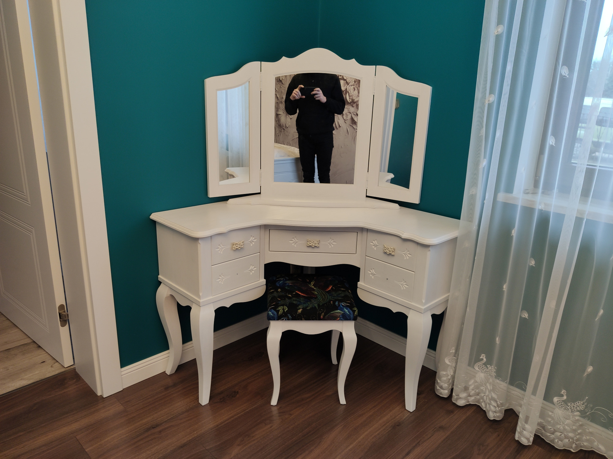 Biała toaletka z trzema lustrami i tapicerowanym stołkiem na tle turkusowej ściany i białych, wzorzystych zasłon, z odbiciem fotografa w lustrze.