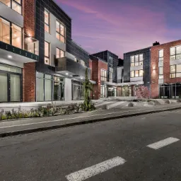 Nowoczesny budynek biurowy z elementami cegły i szarości, oświetlony wieczornym światłem, z widoczną drogą i chodnikiem.