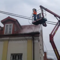 Pracownik w pomarańczowej kamizelce, w koszu podnośnika, czyści dach budynku strumieniem wody pod ciśnieniem; widoczne zacieki i para wodna.