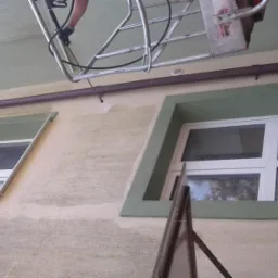 Prace na wysokości: mycie elewacji budynku z podnośnika koszowego, widoczne ślady czyszczenia na jasnej elewacji wokół okien z zielonymi ramami.