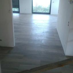 dragon - Jasnoszare płytki podłogowe imitujące drewno, ułożone w nowym budynku, widoczne fragmenty ścian w stanie surowym i drewniane podpory.