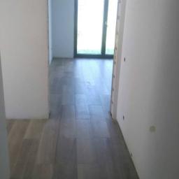 dragon - Korytarz w trakcie remontu z ułożonymi szarymi panelami podłogowymi imitującymi drewno, surowe białe ściany i widok na okno balkonowe w głębi.