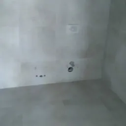 Szare płytki ceramiczne na ścianach i podłodze w łazience, widoczne otwory na instalacje wodno-kanalizacyjne, brak fugowania.