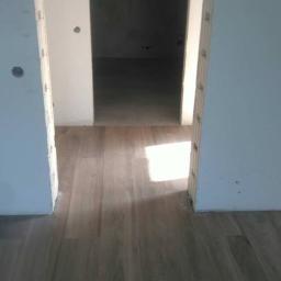 dragon - Wykończona podłoga z paneli imitujących drewno, widok przez futrynę, ściany w trakcie przygotowania do malowania z widocznymi otworami po instalacjach.