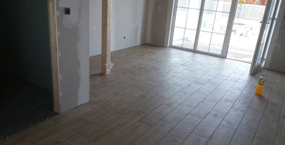 Wyłożona płytkami podłoga z efektem drewna w nowym pomieszczeniu, widoczne niedoskonałości i resztki po pracach, butelka soku w rogu przy drzwiach balkonowych.