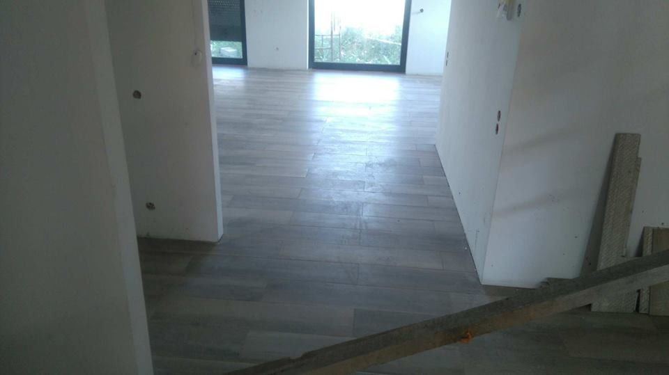 Jasnoszare płytki podłogowe imitujące drewno, ułożone w nowym budynku, widoczne fragmenty ścian w stanie surowym i drewniane podpory.