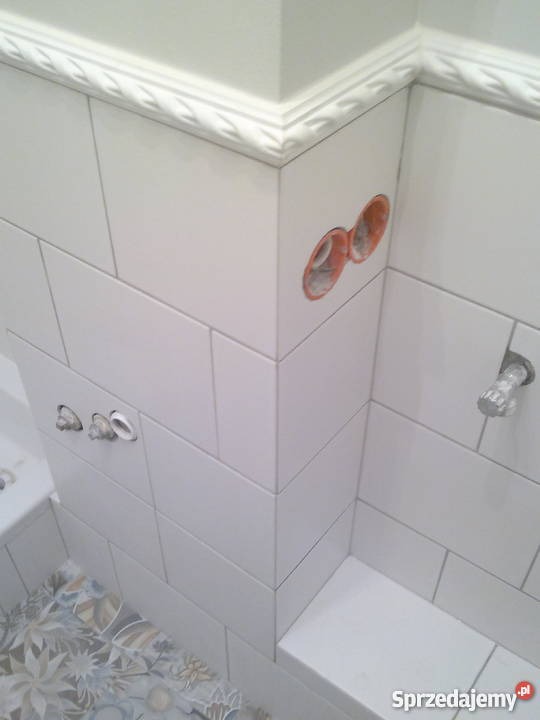 Ściana łazienki wyłożona białymi, prostokątnymi płytkami, ułożonymi w cegiełkę, z widocznymi otworami na instalacje i dekoracyjnym gzymsem pod sufitem.