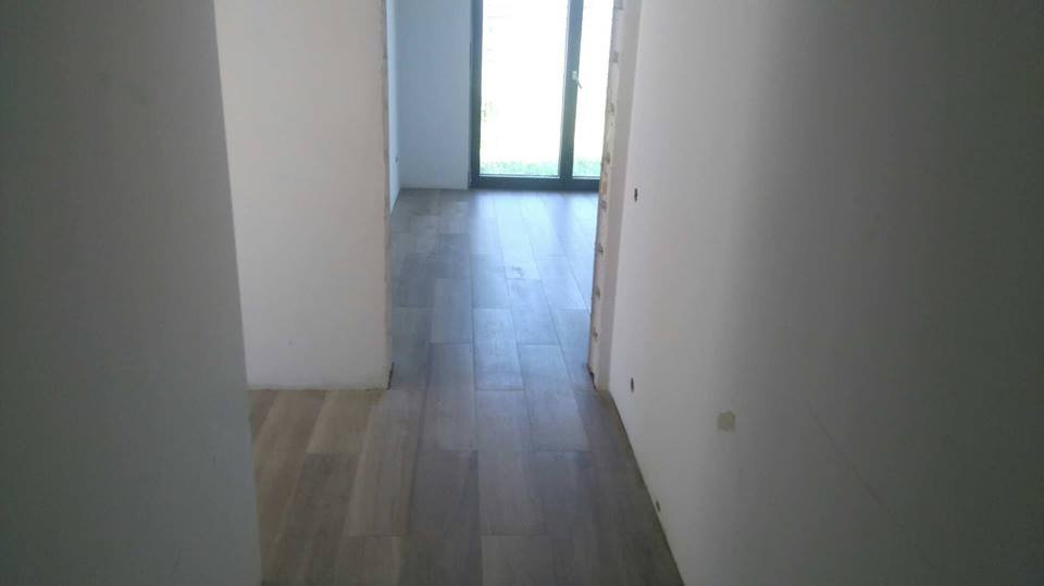 Korytarz w trakcie remontu z ułożonymi szarymi panelami podłogowymi imitującymi drewno, surowe białe ściany i widok na okno balkonowe w głębi.