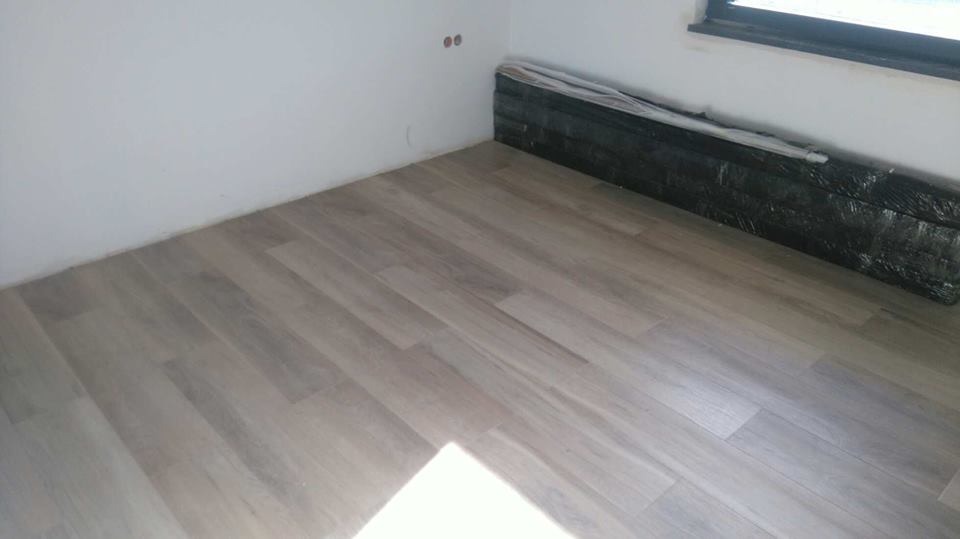 Wykończona podłoga z paneli w odcieniach szarości, ułożona w jasnym pokoju. Zabezpieczone panele czekają na montaż w rogu. Widoczne dwa otwory w ścianie.