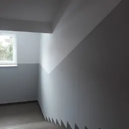 Klatka schodowa z pomalowanymi ścianami na dwa odcienie szarości, widoczne schody wyłożone płytkami i okno z widokiem na zieleń.