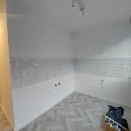 Świeżo wykafelkowana ściana w kuchni z widocznymi gniazdkami elektrycznymi i przyłączami hydraulicznymi, podłoga wyłożona panelami ułożonymi w jodełkę, karton z odpadami w rogu.