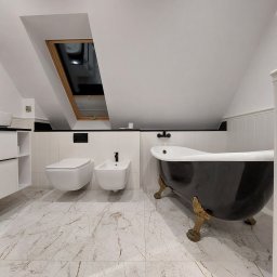 SOLIDNE WNĘTRZA - TOMASZ KIJOWSKI - Nowoczesna łazienka na poddaszu z wolnostojącą czarną wanną na złotych nóżkach, wiszącą toaletą i bidetem z czarną armaturą, marmurową posadzką i oknem dachowym.