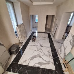 SOLIDNE WNĘTRZA - TOMASZ KIJOWSKI - Układanie płytek podłogowych imitujących marmur w trakcie remontu; widoczny wózek do fugowania, płyty do docinania i surowe ściany.