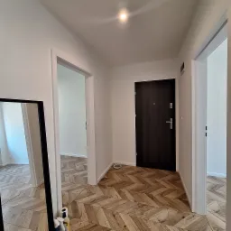 Korytarz po remoncie z podłogą ułożoną w jodełkę z drewnopodobnych paneli, białe ściany i drzwi, widoczne przejścia do innych pomieszczeń, w rogu przy lustrze leżą środki czystości.