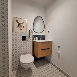 SOLIDNE WNĘTRZA - TOMASZ KIJOWSKI - Nowoczesna łazienka z wiszącą szafką z drewnianymi frontami, owalnym lustrem w czarnej ramie, toaletą i płytkami w geometryczny wzór na podłodze i ścianie za toaletą.