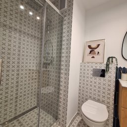 SOLIDNE WNĘTRZA - TOMASZ KIJOWSKI - Nowoczesna łazienka z kabiną prysznicową typu walk-in, wykończona płytkami w geometryczny wzór, wisząca toaleta z chromowanym przyciskiem, ozdobny obrazek i roślina w dekoracyjnej doniczce.
