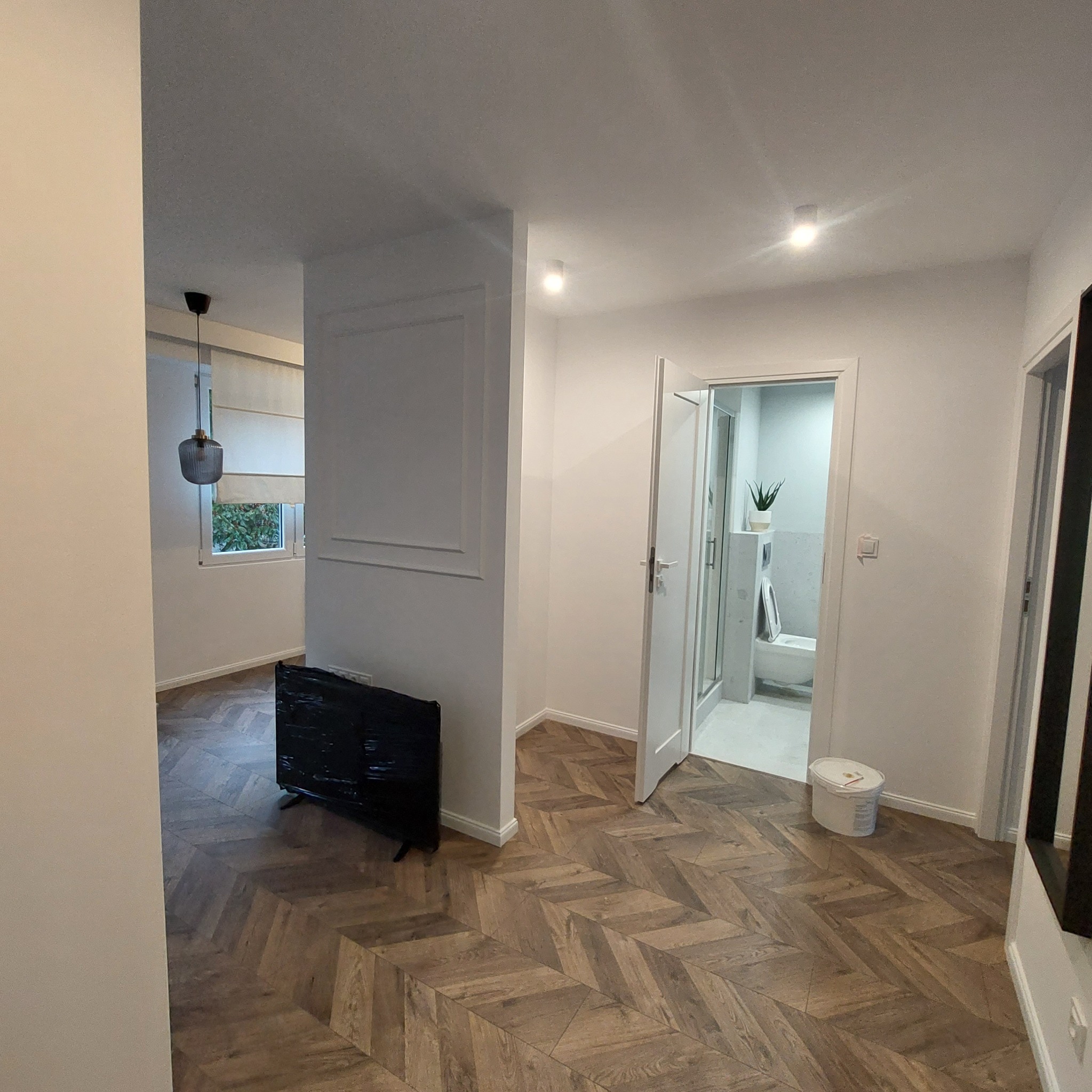 Nowoczesne wnętrze po renowacji: parkiet ułożony we wzór jodełki, białe ściany z dekoracyjnymi listwami, widok na odnowioną łazienkę z kabiną prysznicową i toaletą, oraz zabezpieczony folią telewizor.