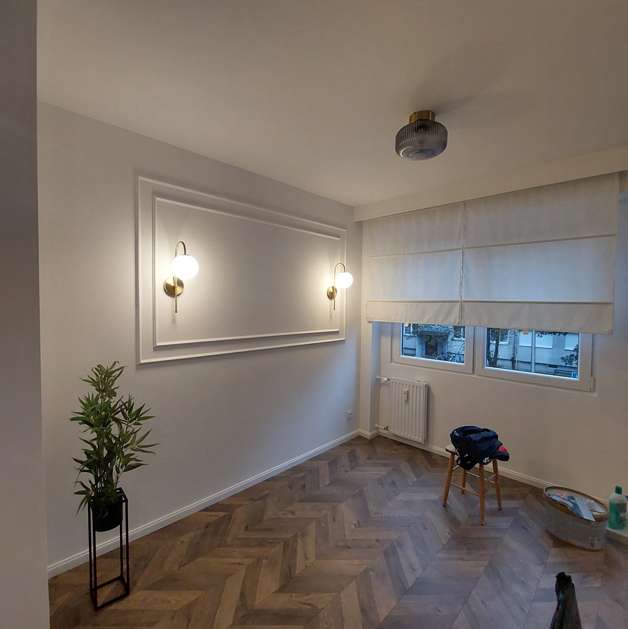 Jasne wnętrze z parkietem w jodełkę, dekoracyjną sztukaterią na ścianie, złotymi kinkietami i roletą rzymską w oknie.