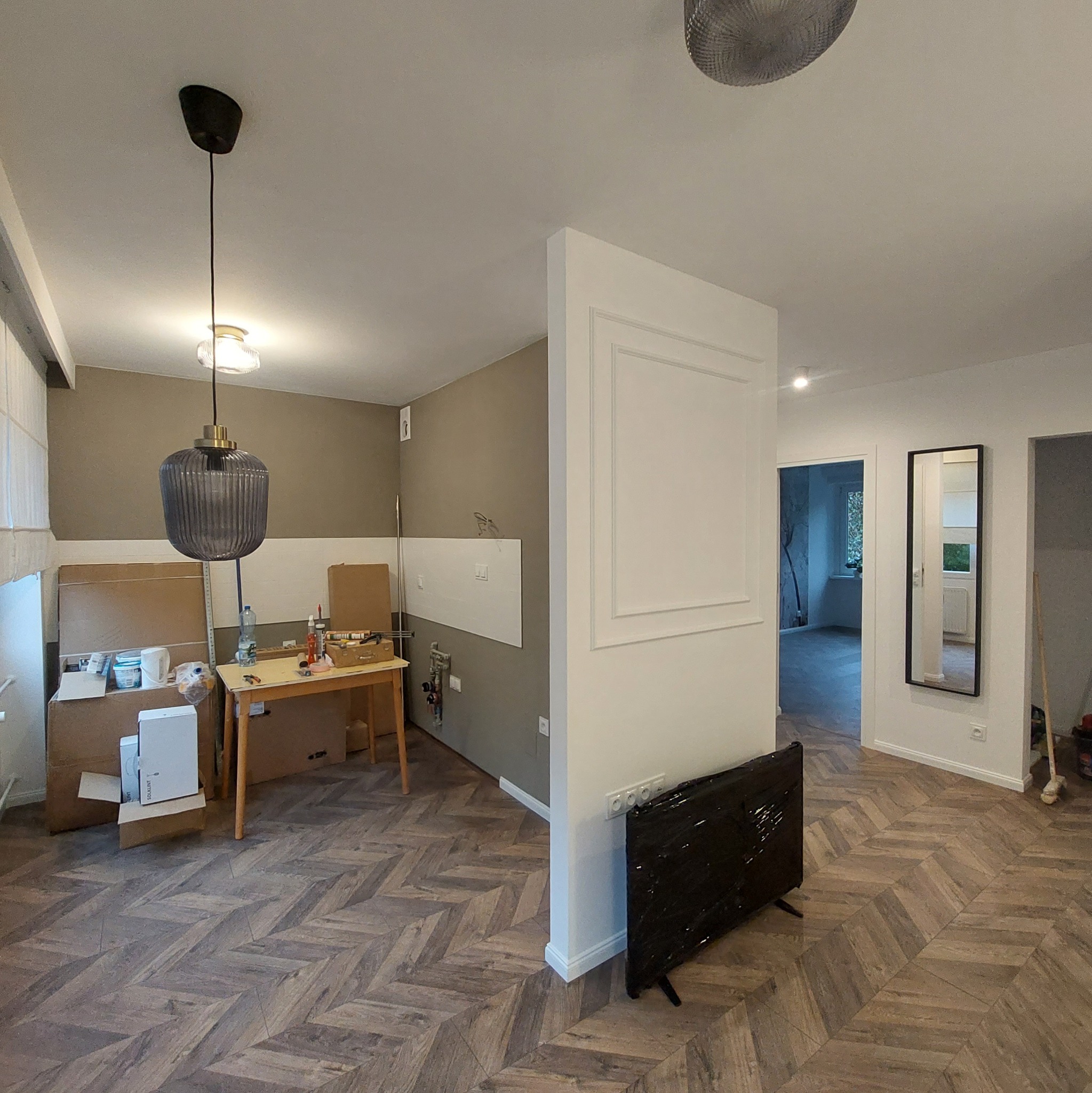 Wnętrze mieszkania w trakcie prac wykończeniowych: widoczne panele ułożone w jodełkę, częściowo pomalowane ściany, kartony z farbami, zabezpieczony grzejnik, wisząca lampa i roboczy stół...