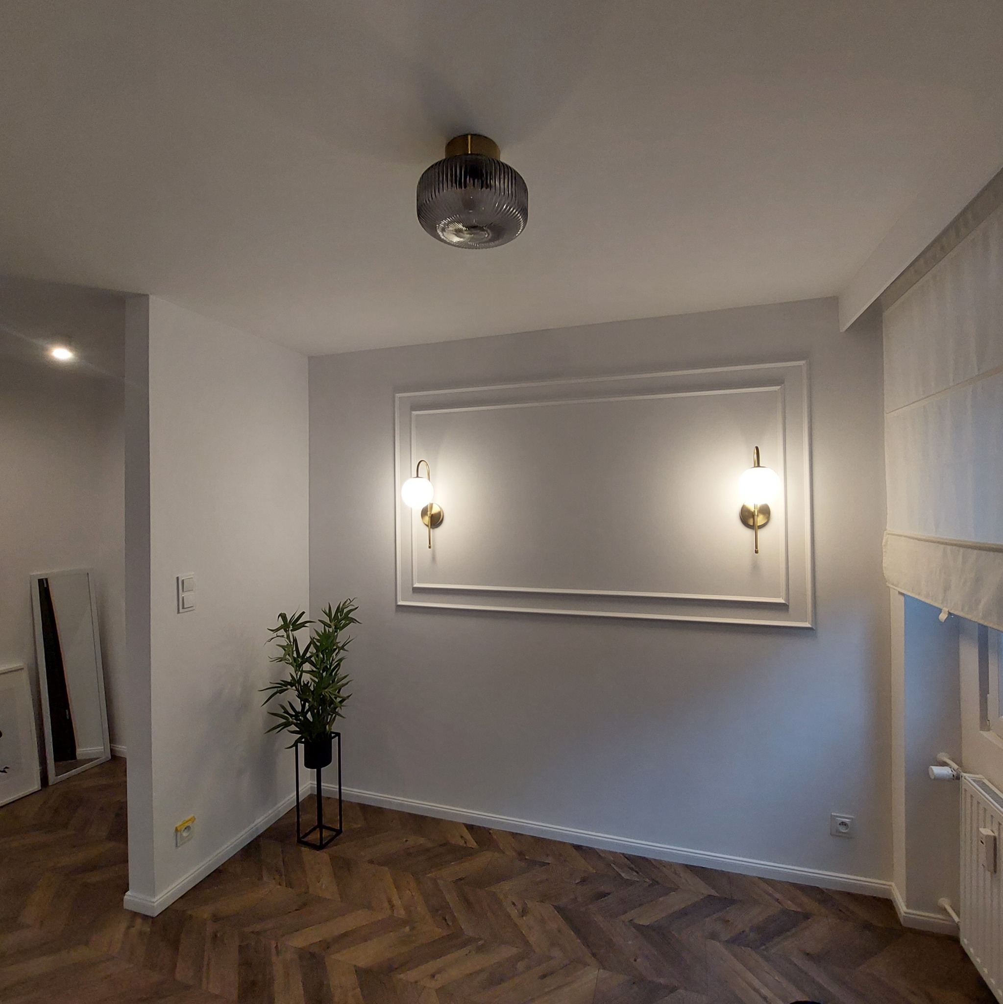 Nowoczesne wnętrze z parkietem w jodełkę, ozdobnymi listwami ściennymi, kinkietami z białymi kulami i minimalistyczną lampą sufitową, uzupełnione zieloną rośliną w czarnej donicy.