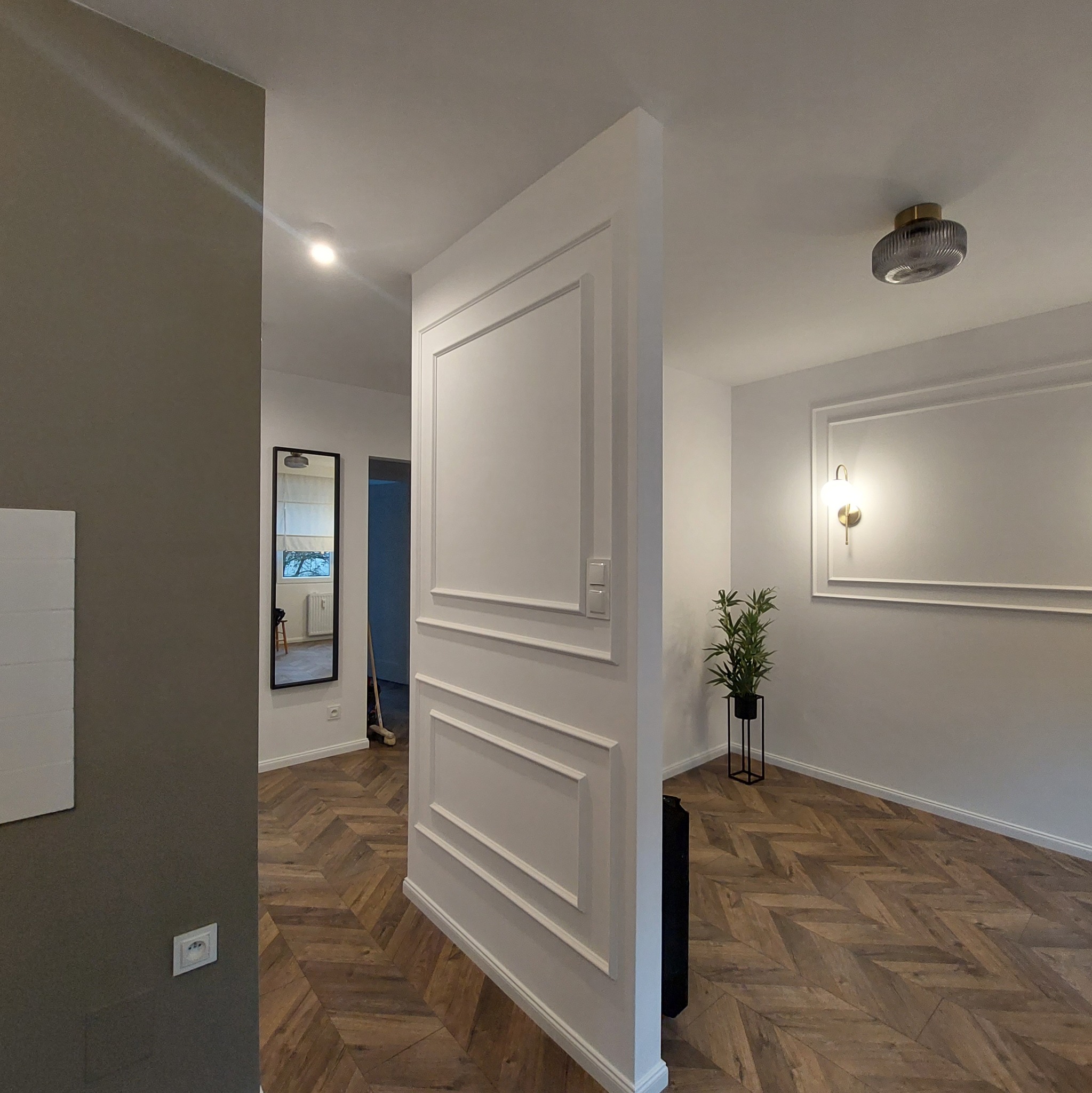 Nowoczesne wnętrze z parkietem w jodełkę, białą ścianką działową z dekoracyjnymi listwami, lustrem w czarnej ramie i rośliną w metalowej donicy.