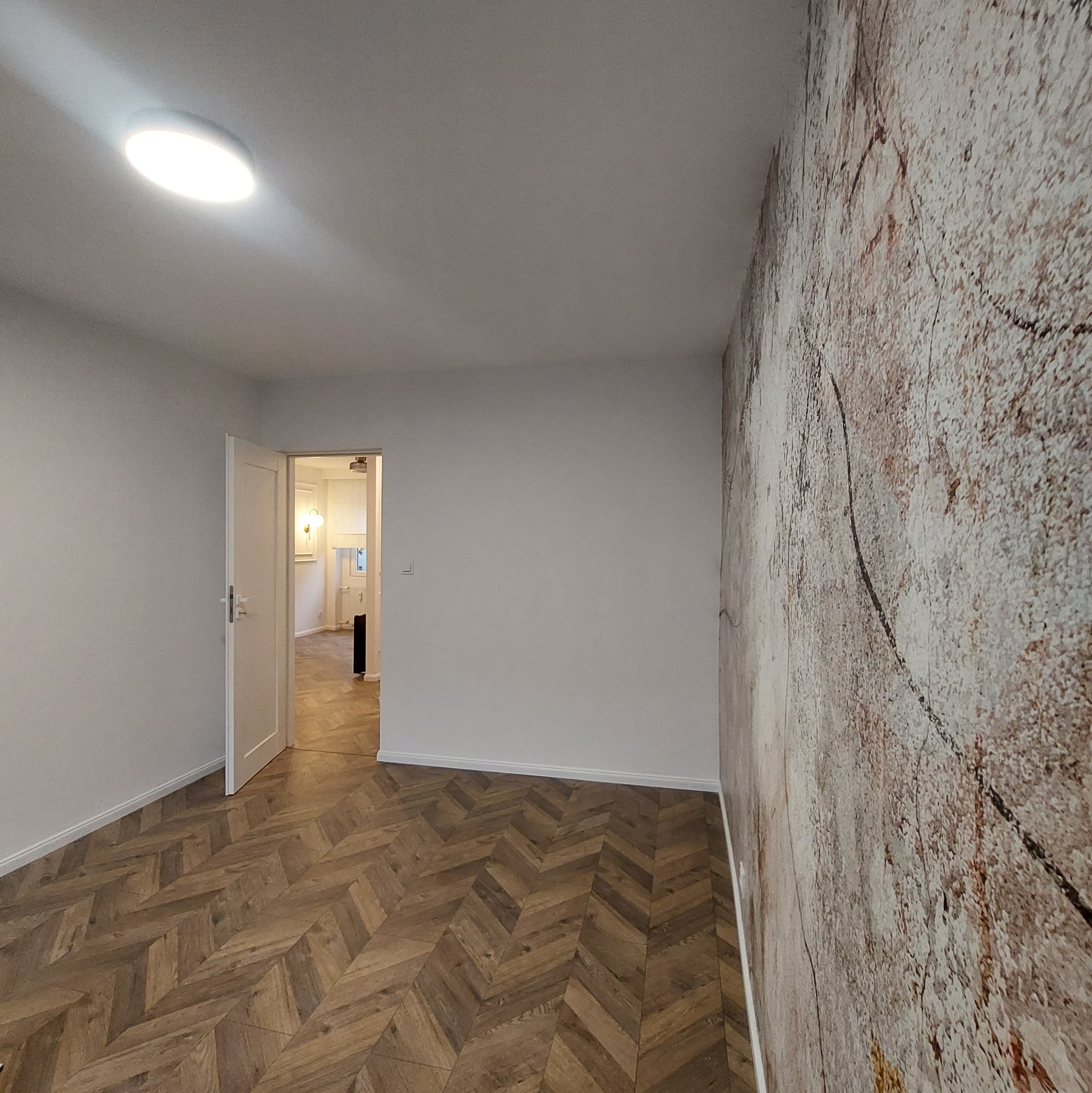 Pusty pokój z drewnianą podłogą ułożoną w jodełkę, białe ściany, jedna ściana pokryta tapetą imitującą kamień i otwarte białe drzwi do kolejnego pokoju.