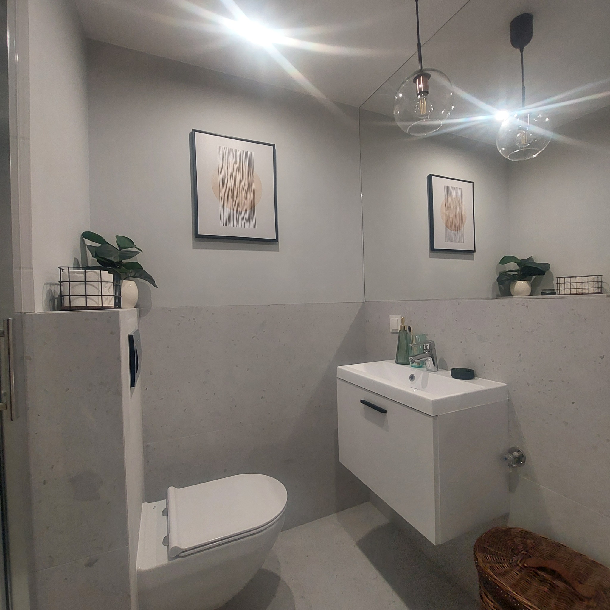 Nowoczesna łazienka z podwieszaną szafką umywalkową, wiszącą toaletą, lustrem, dekoracyjnym obrazem i rośliną doniczkową, oświetlona dwoma szklanymi lampami.